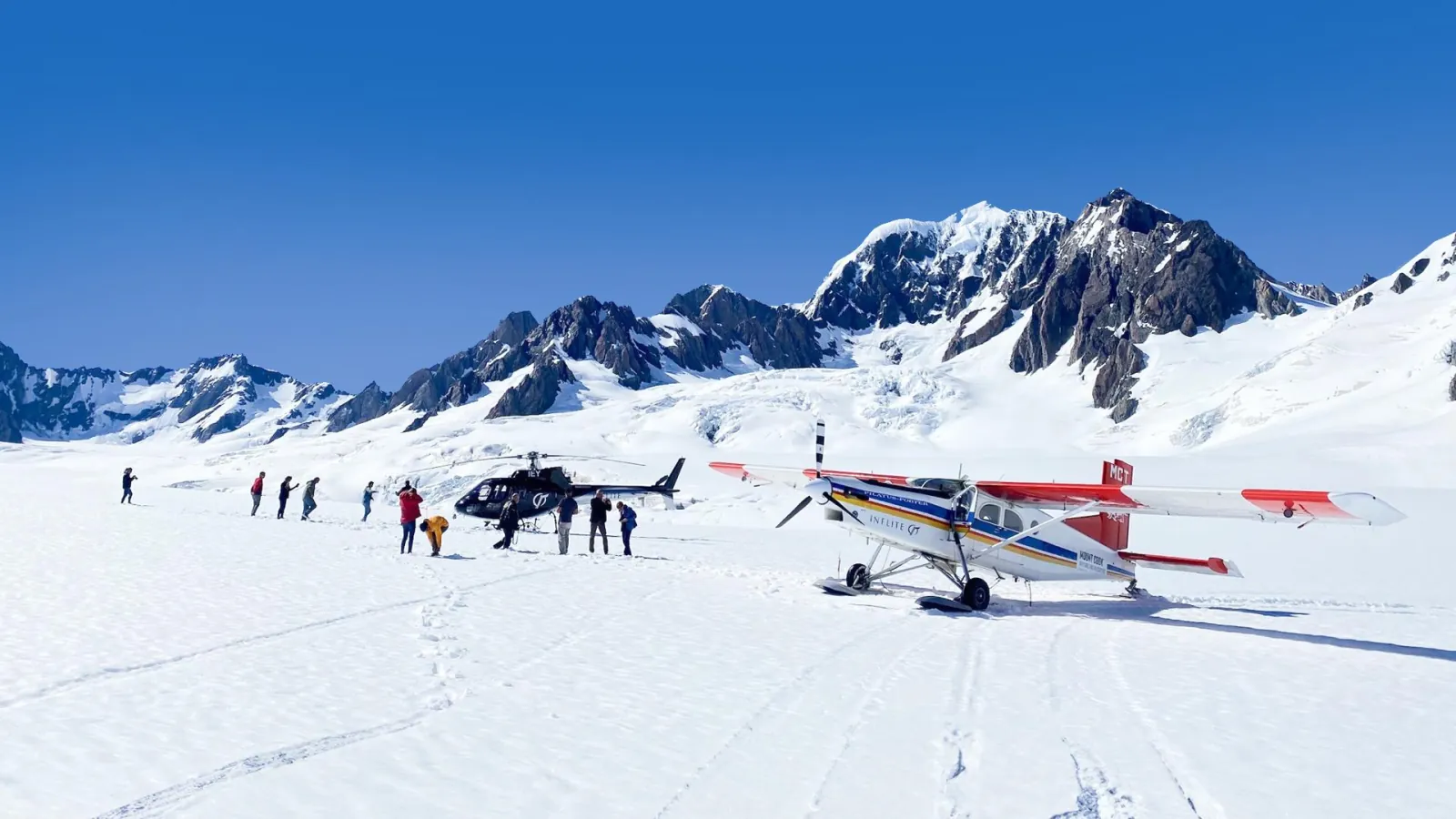 Pilatus Porter ski plane and a black helicopter on a high snowfield, passengers walking between them