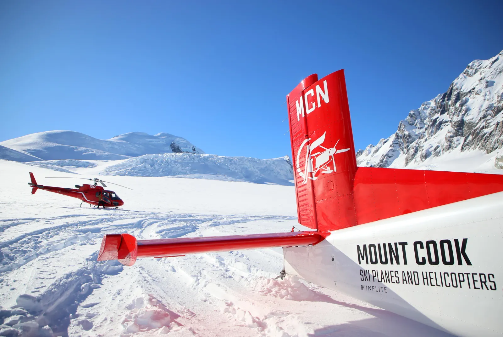 Tail of a Mount Cook Ski Planes aircraft on the glacier snow, a red helicopter parked beyond
