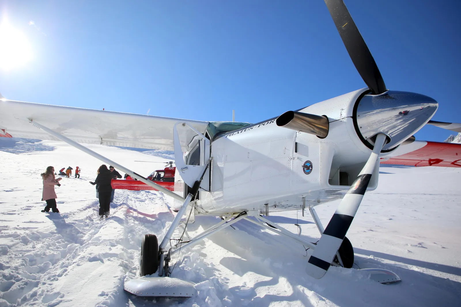 Close-up of a Pilatus Porter ski plane's propeller and skis on the snow, passengers and a red helicopter behind