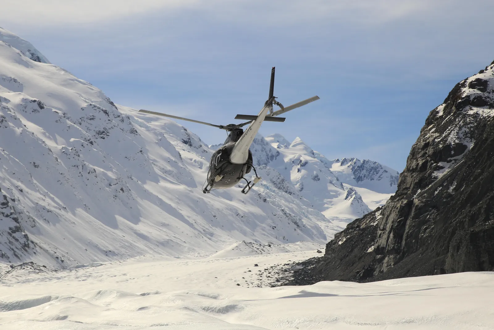 Black helicopter taking off from a glacier snowfield toward snow-covered peaks