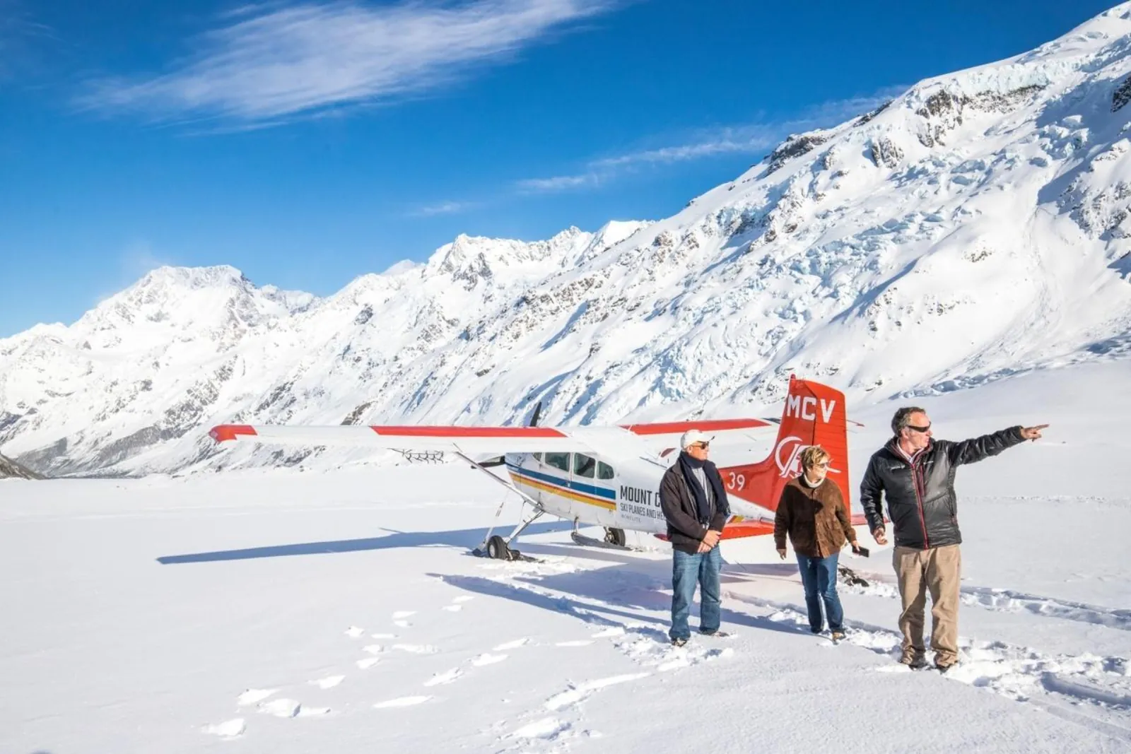 Pilot pointing out peaks to two passengers beside a ski plane on the snow of the Tasman Glacier
