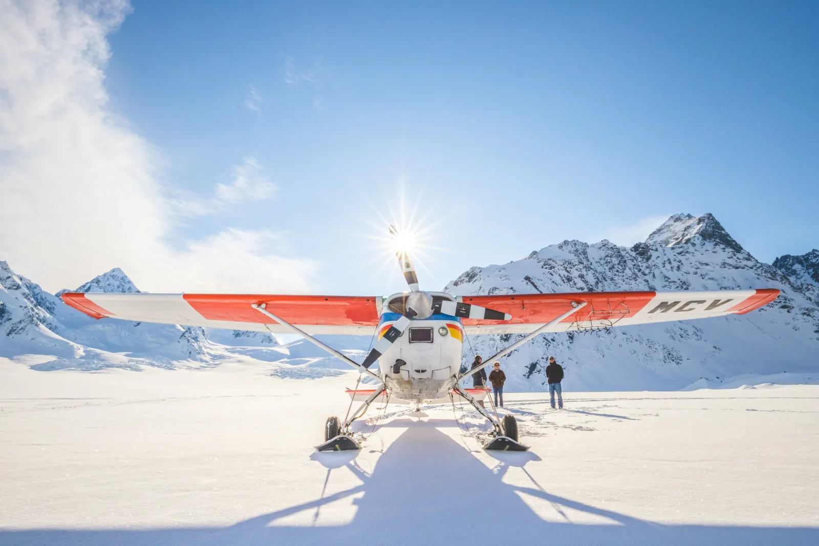 Front view of a red and white ski plane on its skis on a glacier snowfield, sun behind the propeller