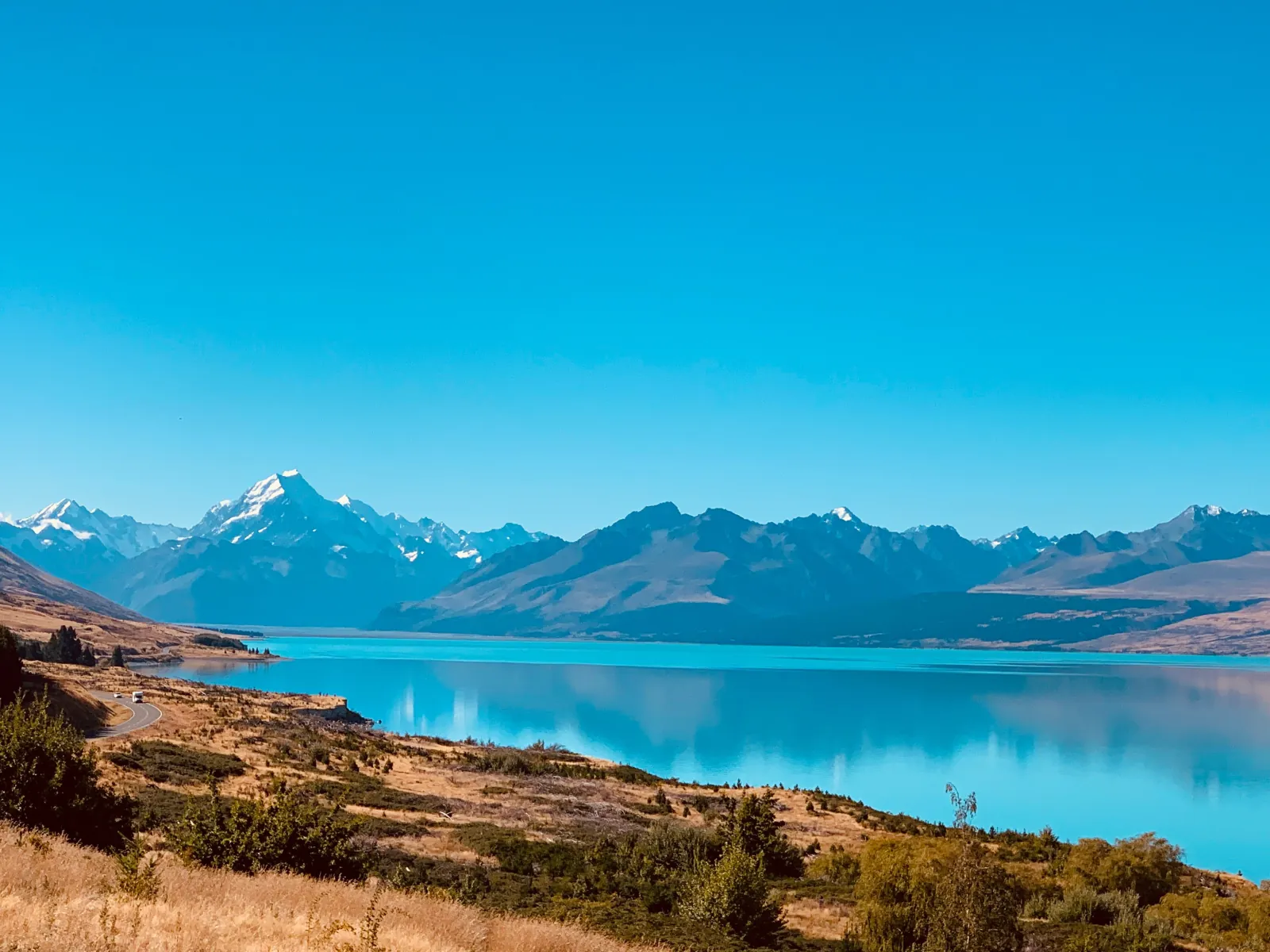 Turquoise Lake Pūkaki under a clear sky, with the road along its shore and Aoraki/Mount Cook on the skyline