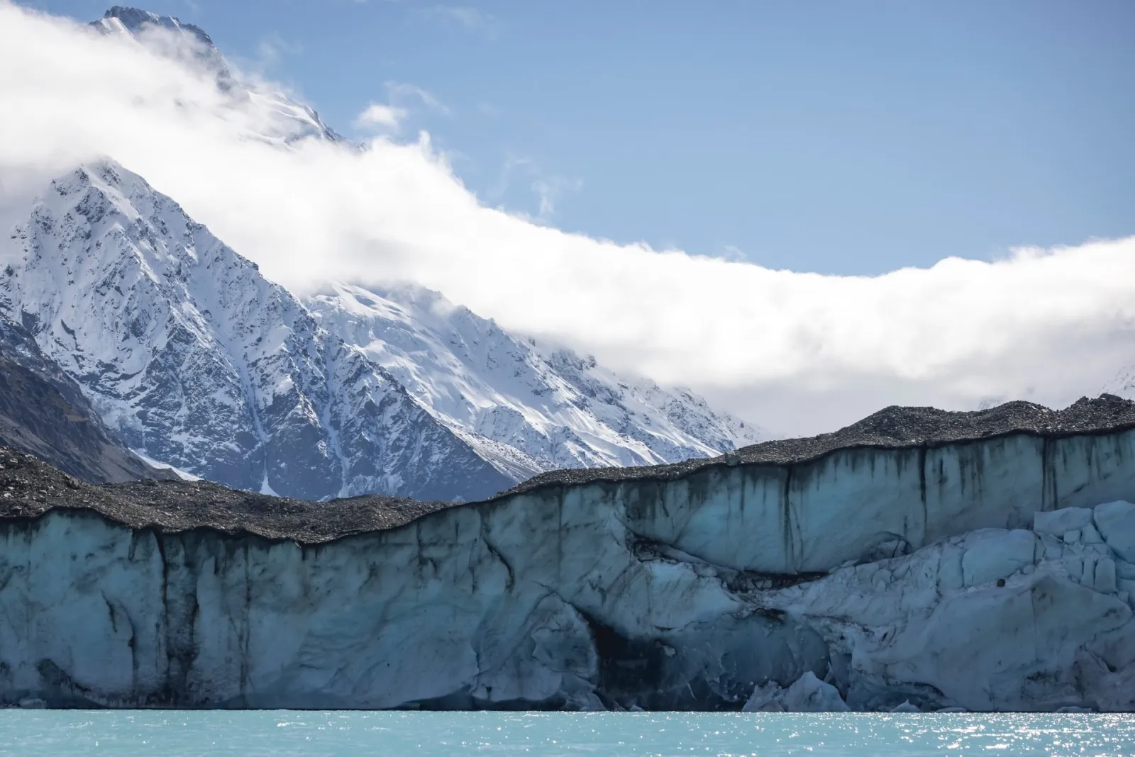 Grey-blue ice cliff of the Tasman Glacier terminal above Tasman Lake, with snowy peaks in cloud behind