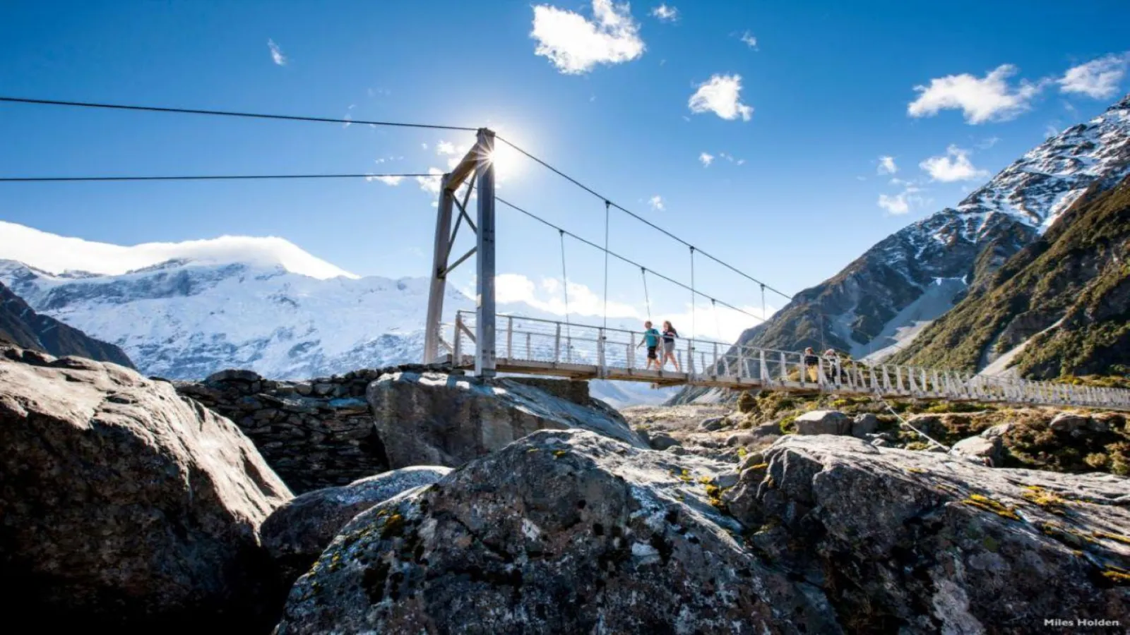 Walkers crossing a swing bridge on the Hooker Valley Track below snowy peaks
