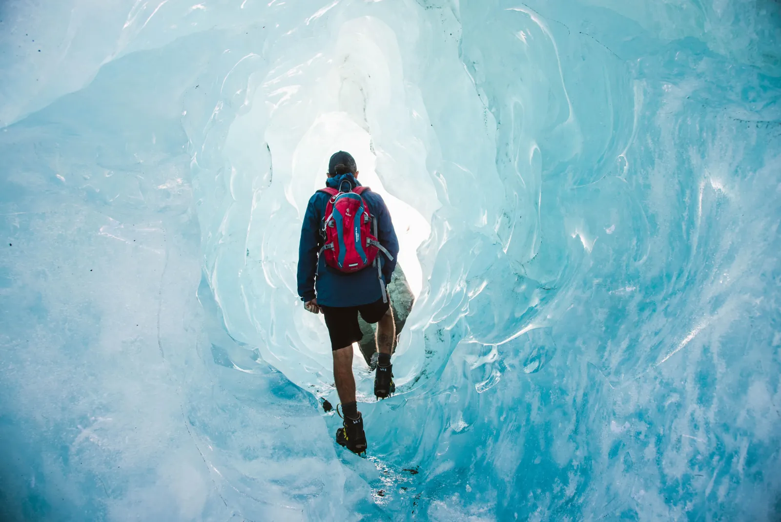 Hiker with a red daypack and crampons walking into a tunnel of blue glacier ice