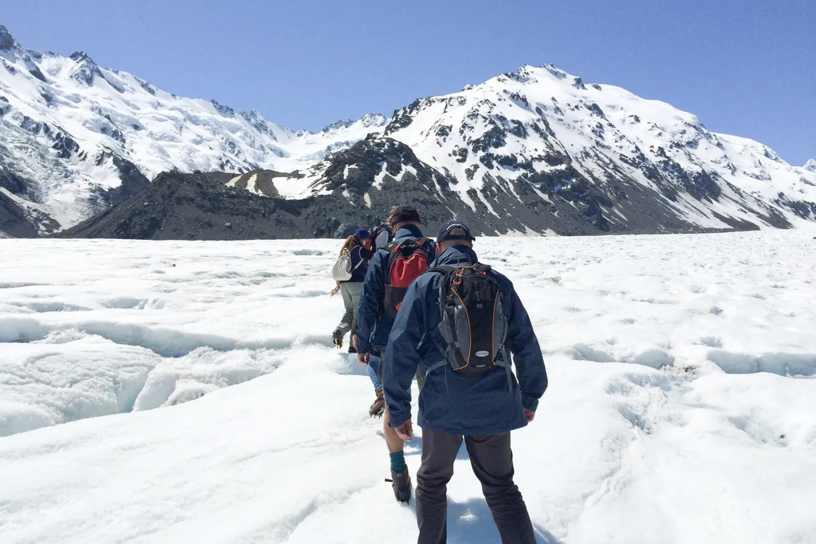 Hikers in single file crossing the white ice of the Tasman Glacier toward snow-covered mountains