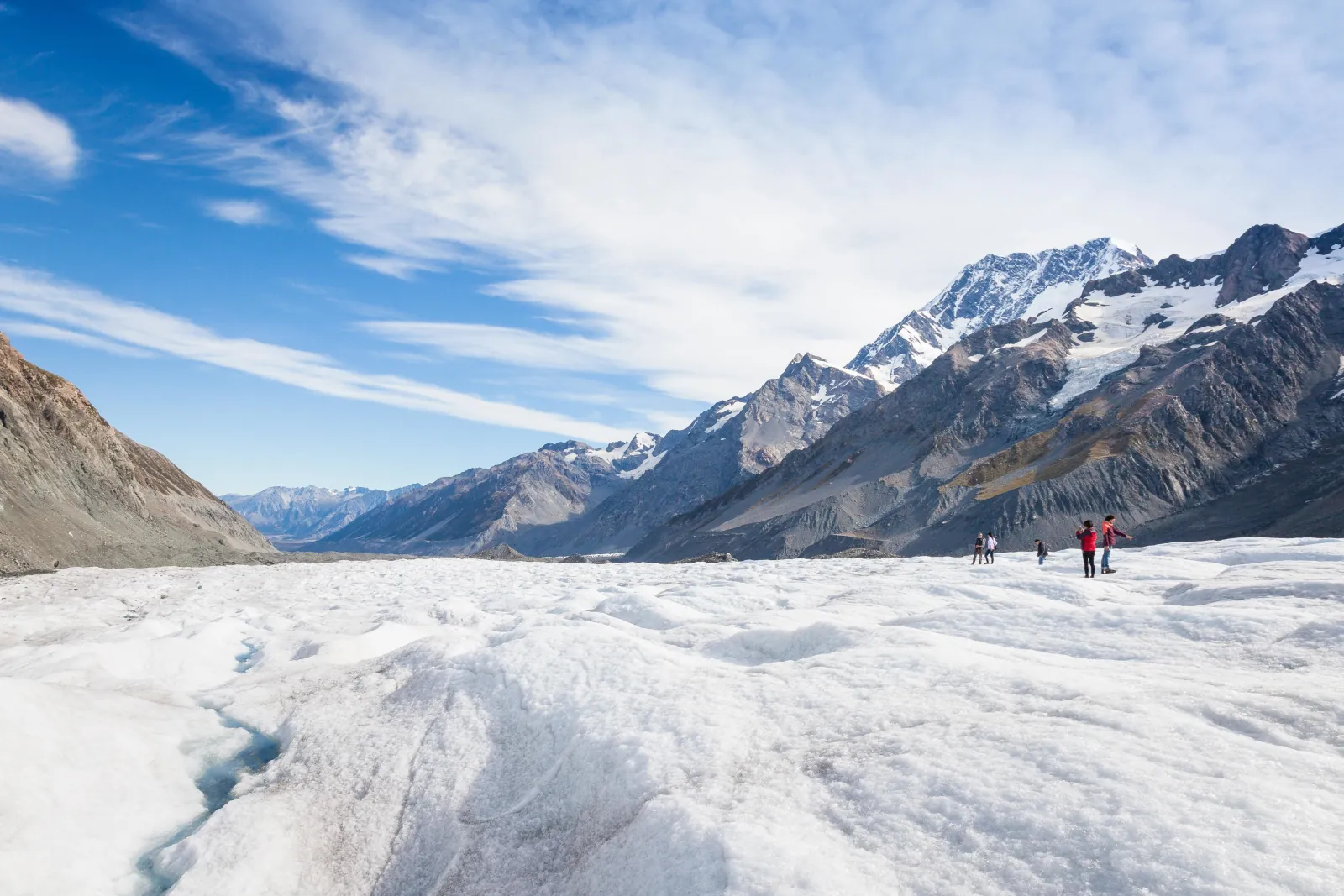 Small group of hikers on the wide ice of the Tasman Glacier below a line of rugged mountains