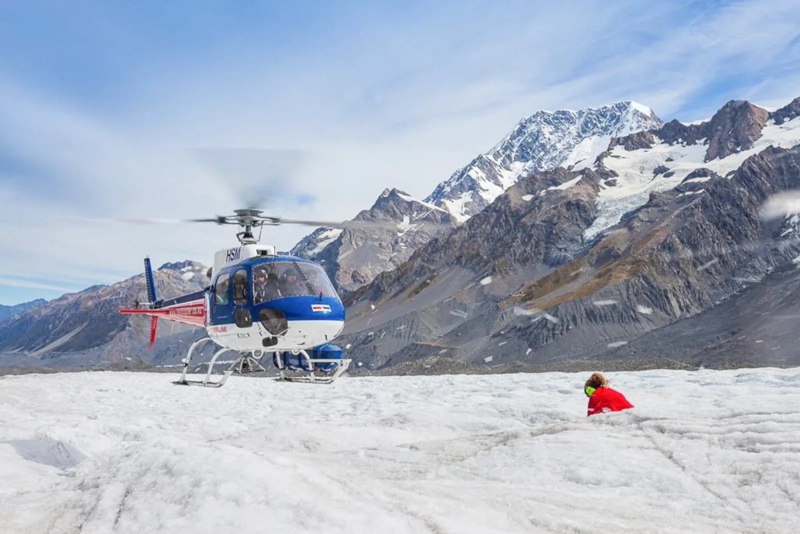 Helicopter with rotors turning on the glacier ice as a hiker in red kneels nearby, snowy peaks behind