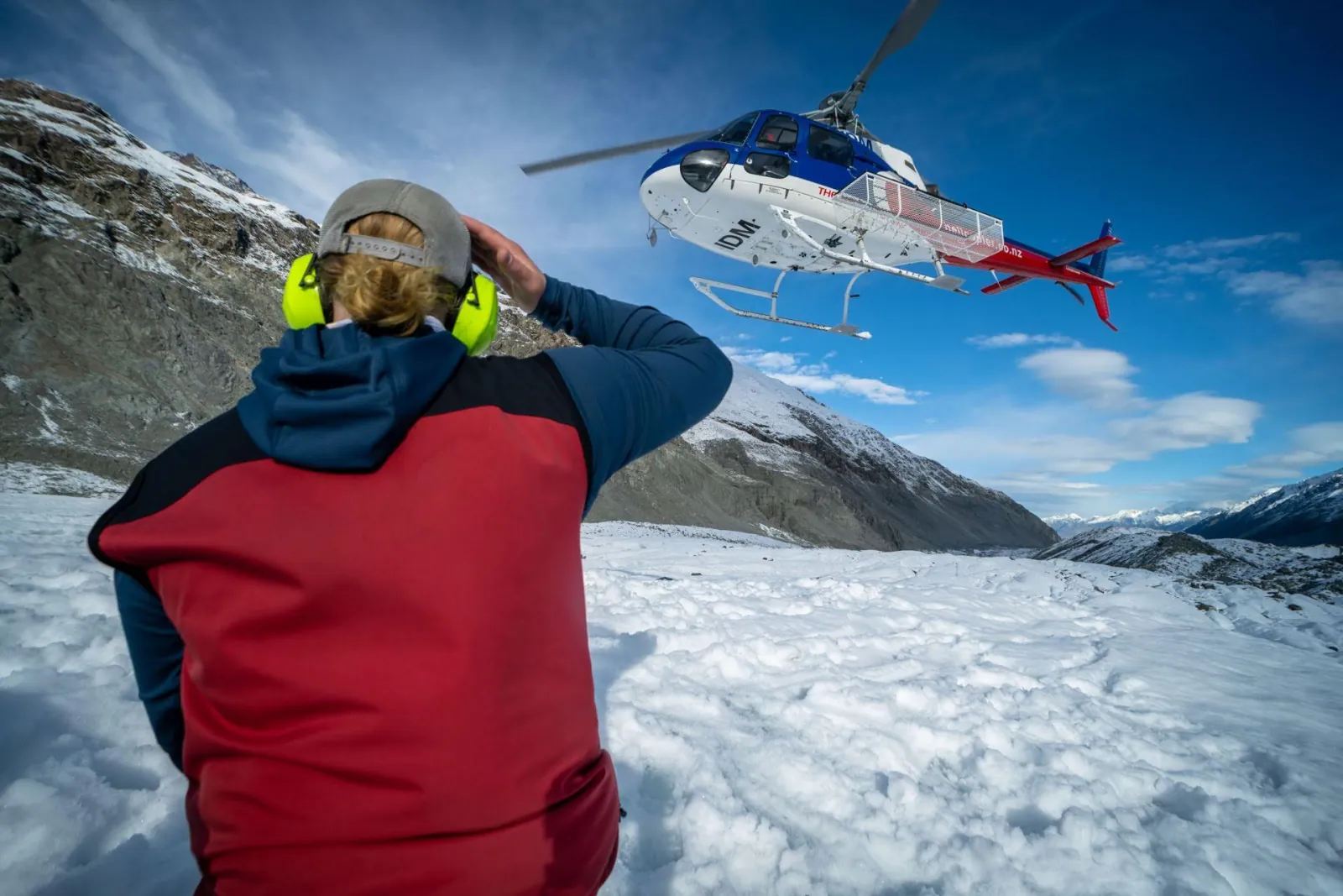 Guide in ear defenders shielding their eyes as a helicopter lifts off from the snowy glacier