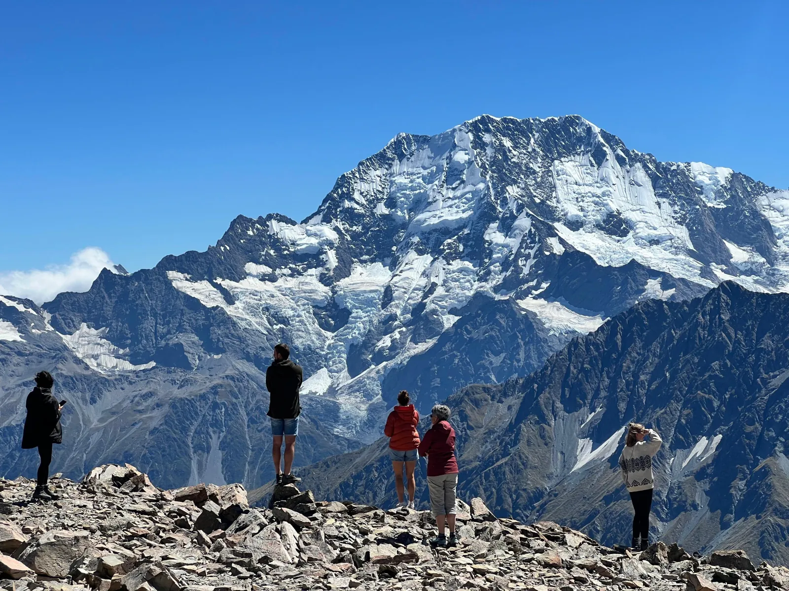 Passengers standing on a rocky alpine landing, looking at a heavily glaciated peak across the valley