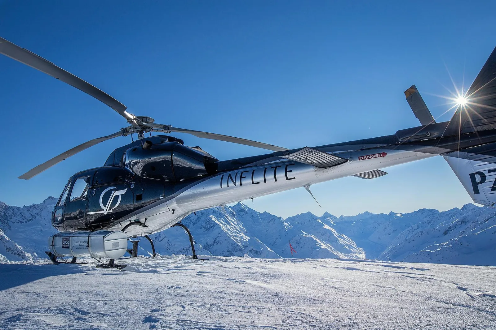 Black and silver INFLITE helicopter on a snowfield in sunshine, snow-covered mountains behind