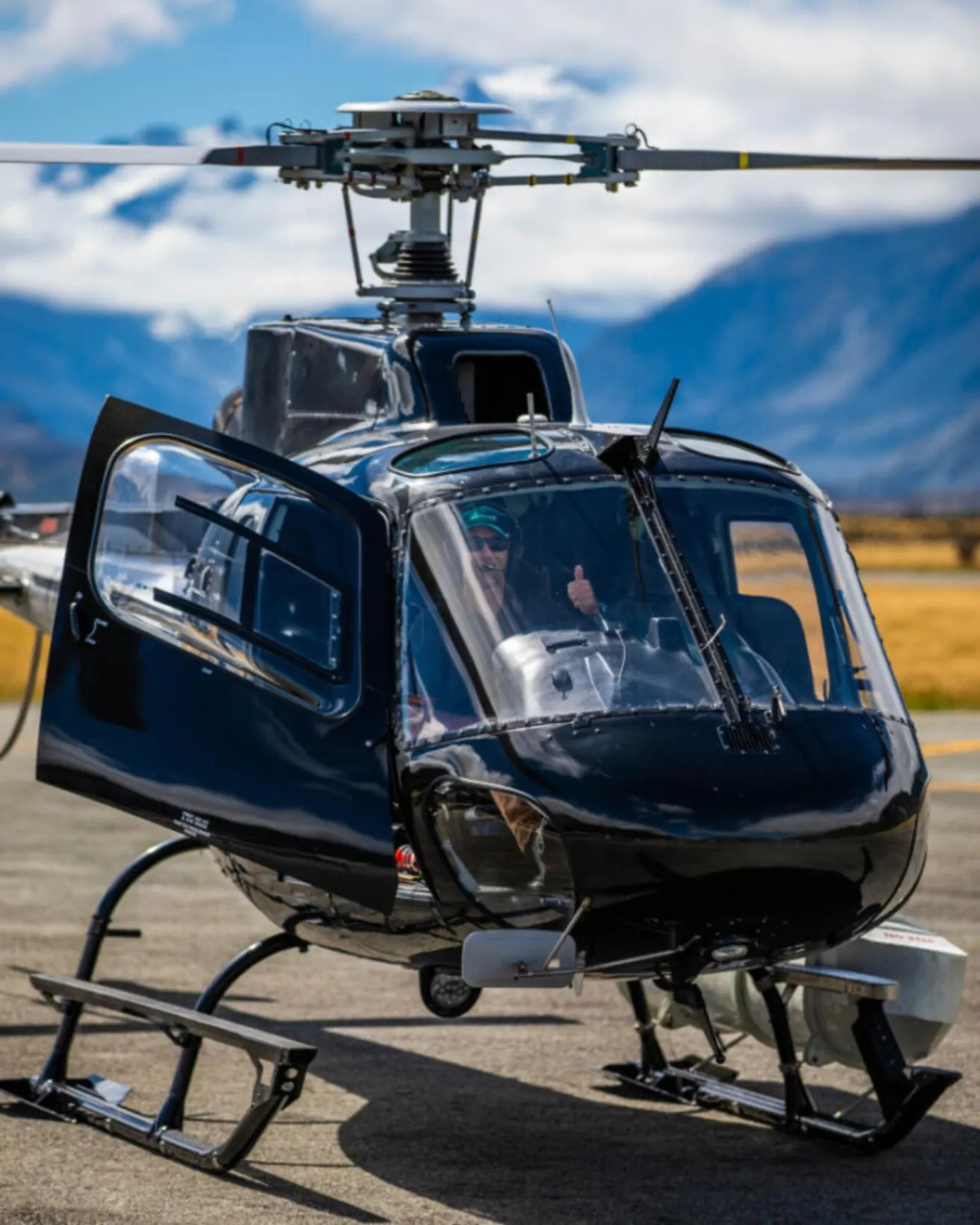 Pilot giving a thumbs up from the cockpit of a black helicopter on the tarmac at Mount Cook Airport