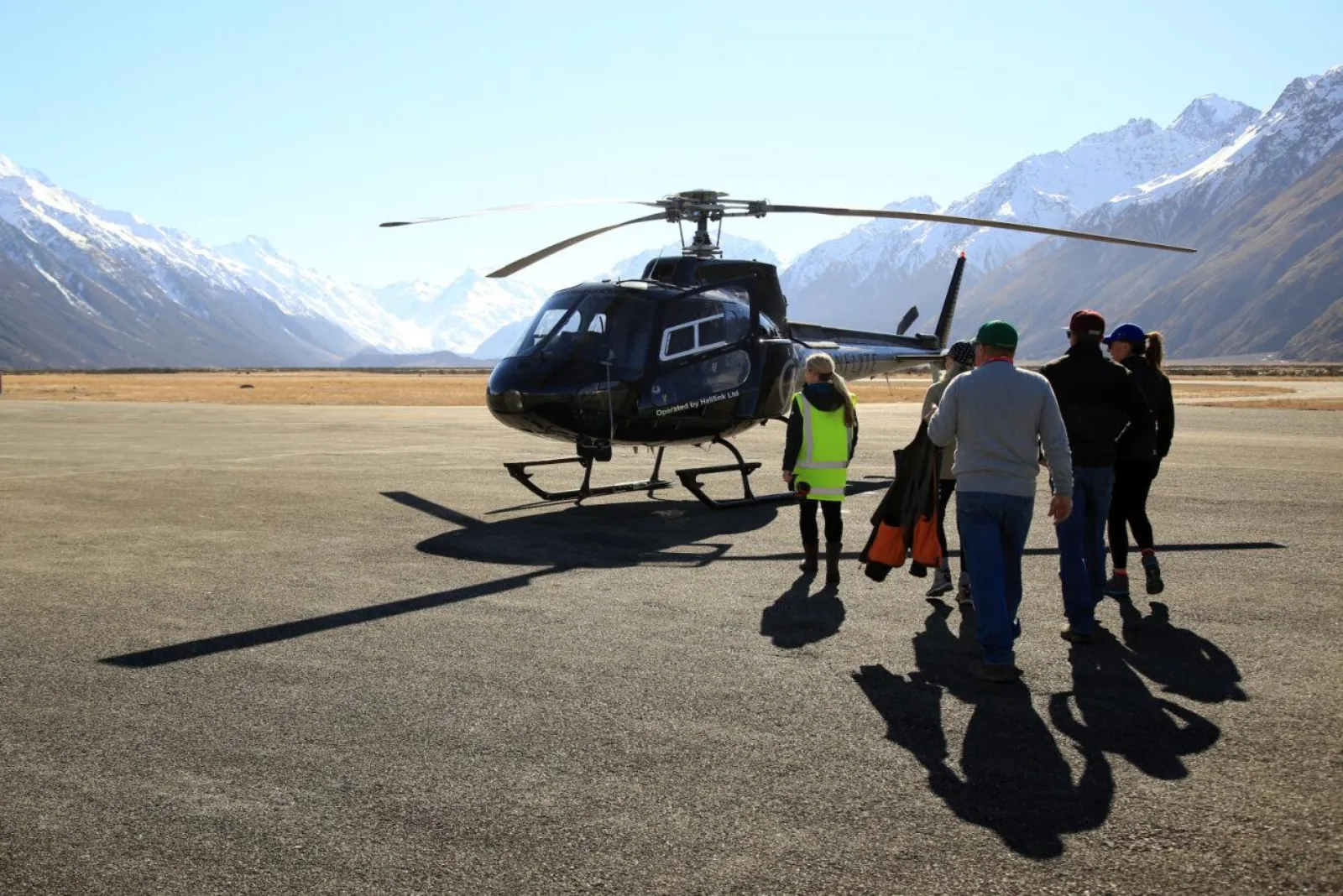 Passengers walking across the apron at Mount Cook Airport to a black helicopter, snowy peaks behind