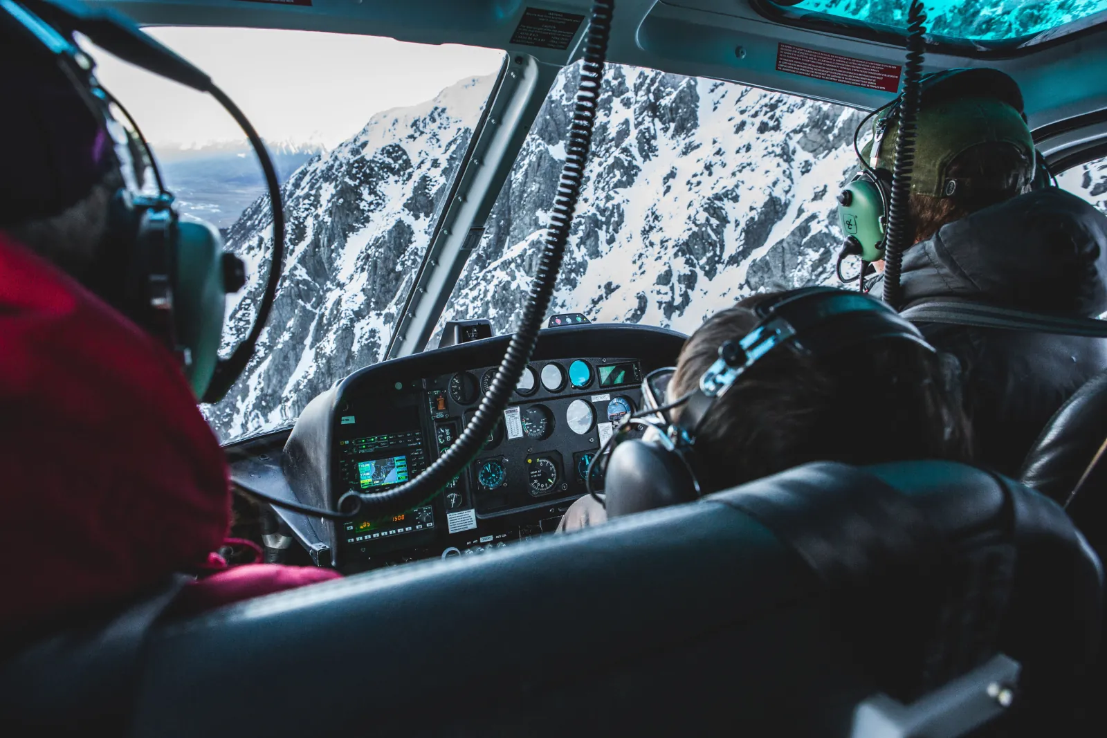 View from a helicopter's rear seat past the pilot and instrument panel to a snowy rock face