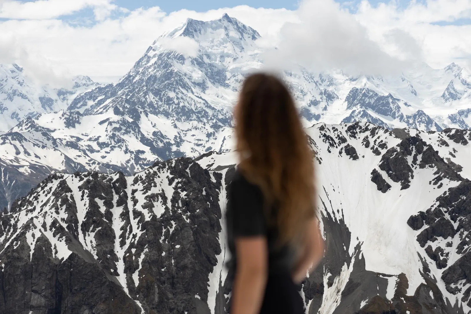A woman, blurred in the foreground, looking at Aoraki/Mount Cook over snow-streaked ridges