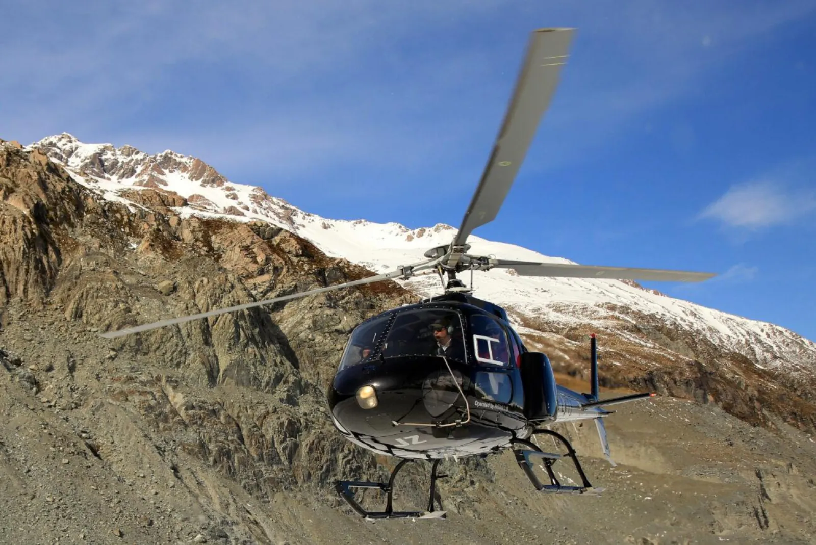 Black helicopter lifting off in front of a rocky moraine slope below a snow-capped ridge