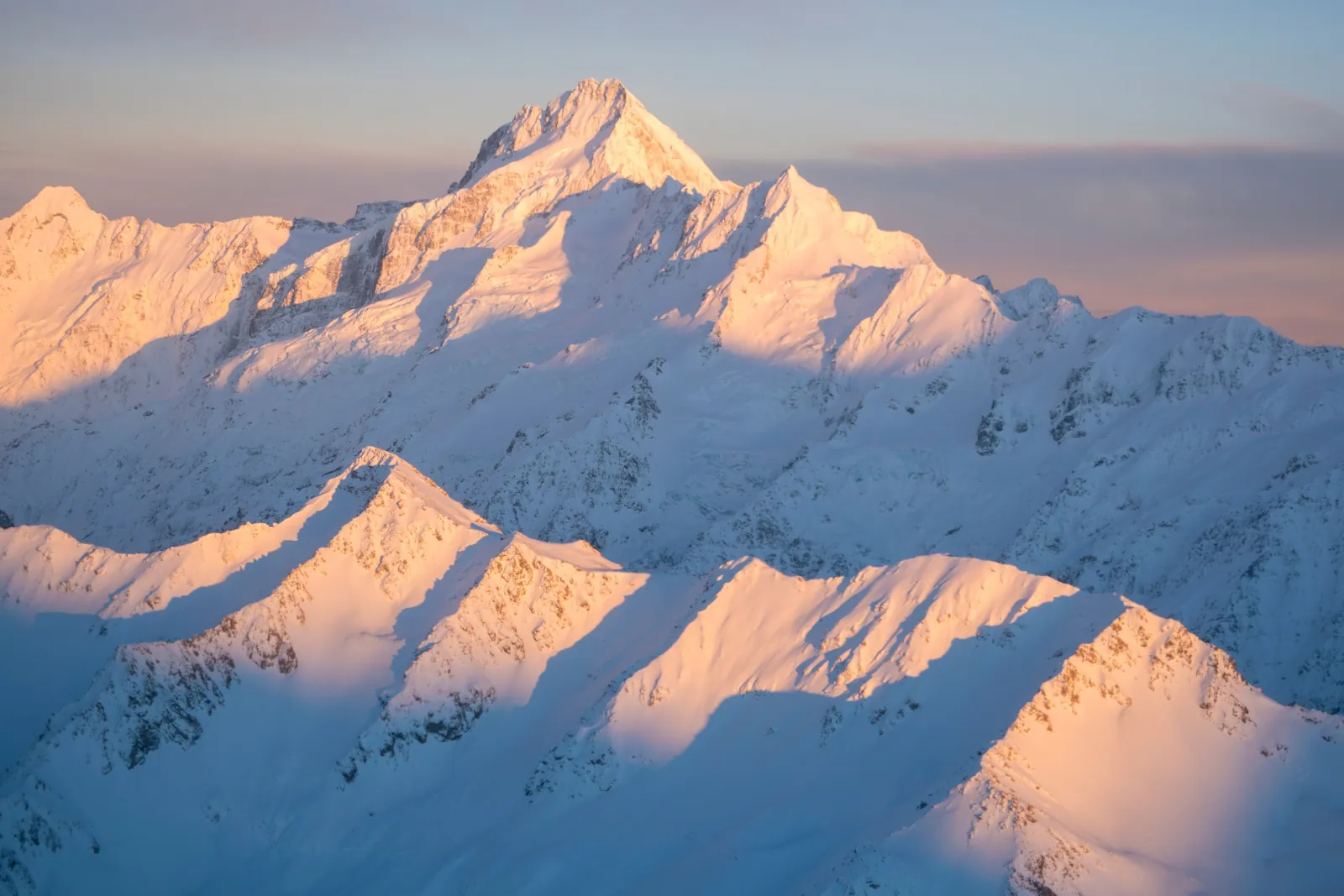 Snow-covered peaks of the Southern Alps lit pink and gold by low sun, seen from the air
