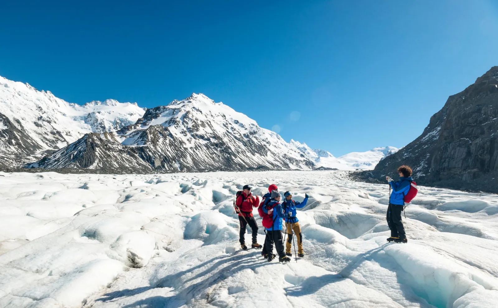 Glacier guide and hikers in crampons on rippled glacier ice below snowy peaks, one hiker taking a photo
