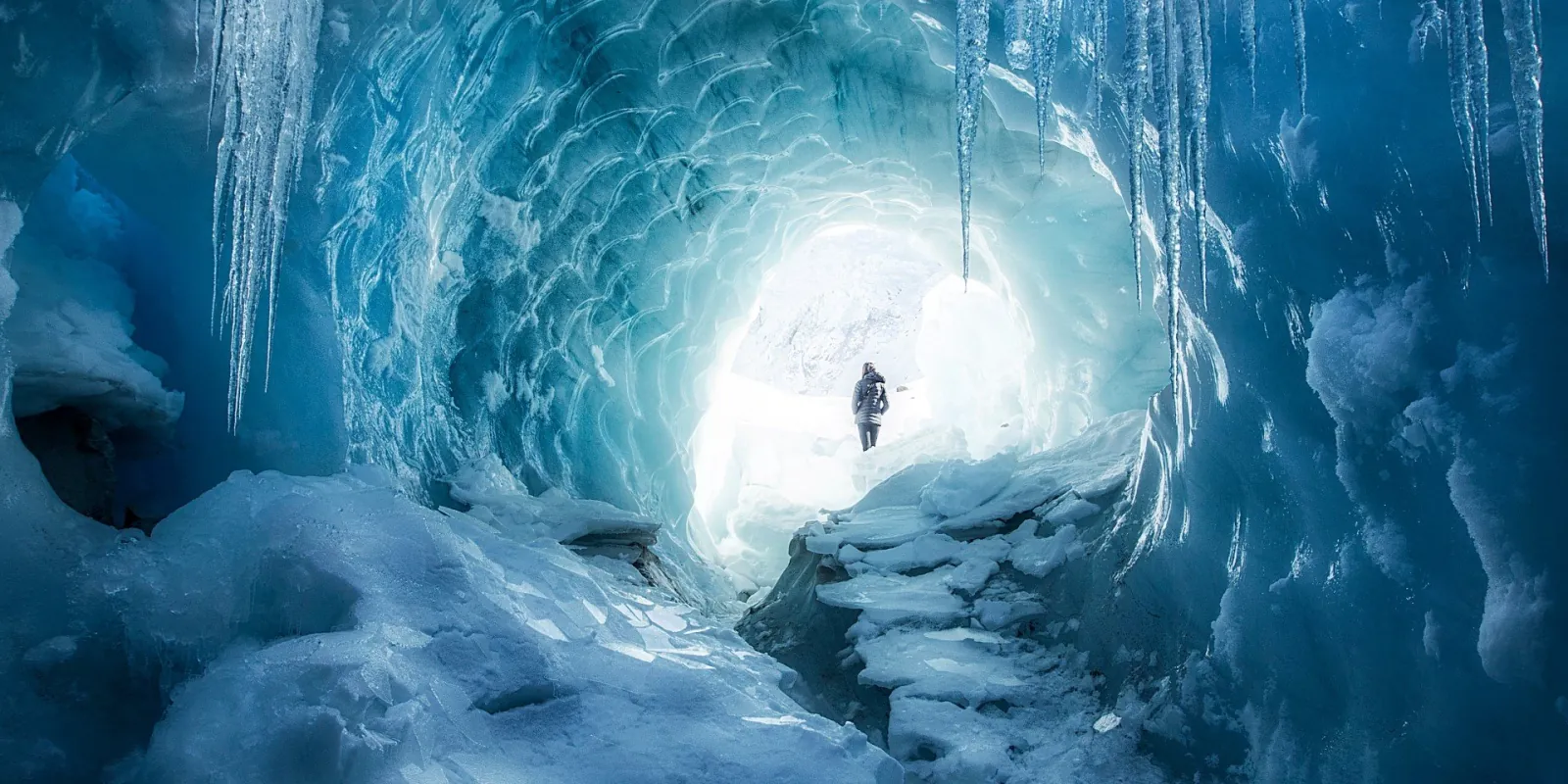 Walker at the bright mouth of a blue ice cave hung with icicles