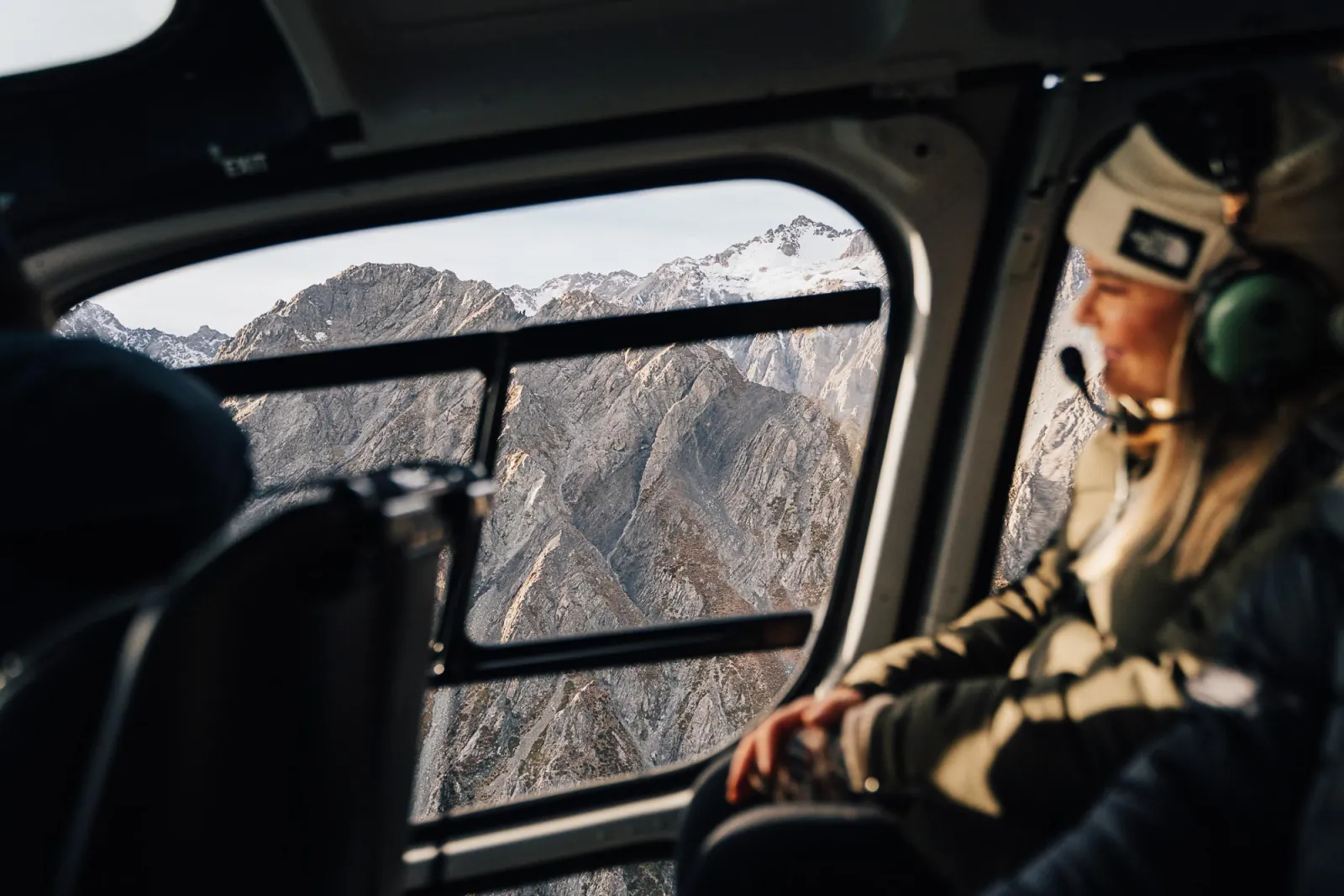 Passenger in a headset looking out of a helicopter window at grey rocky peaks topped with snow