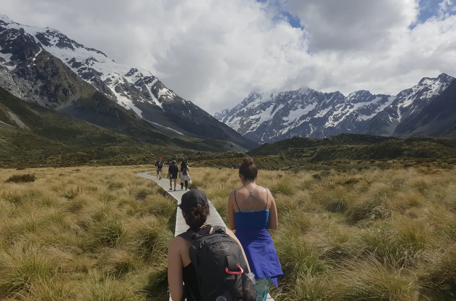 Walkers seen from behind on the Hooker Valley boardwalk, with snow-streaked peaks and cloud ahead