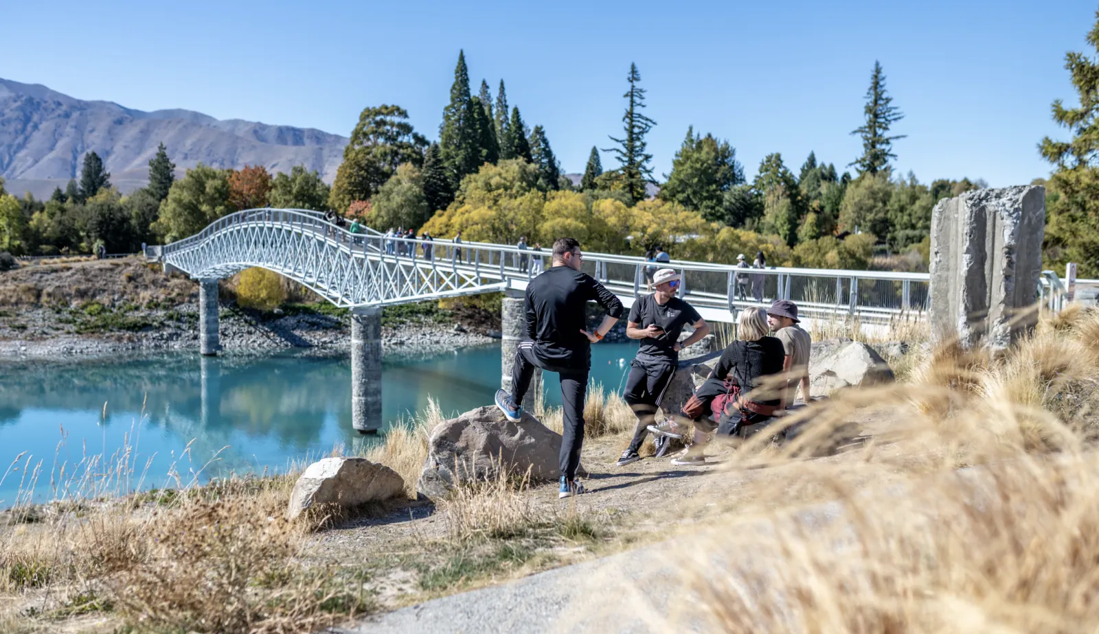 Travelers chatting on the shore of Lake Tekapo beside the white footbridge over the outlet, in bright sun