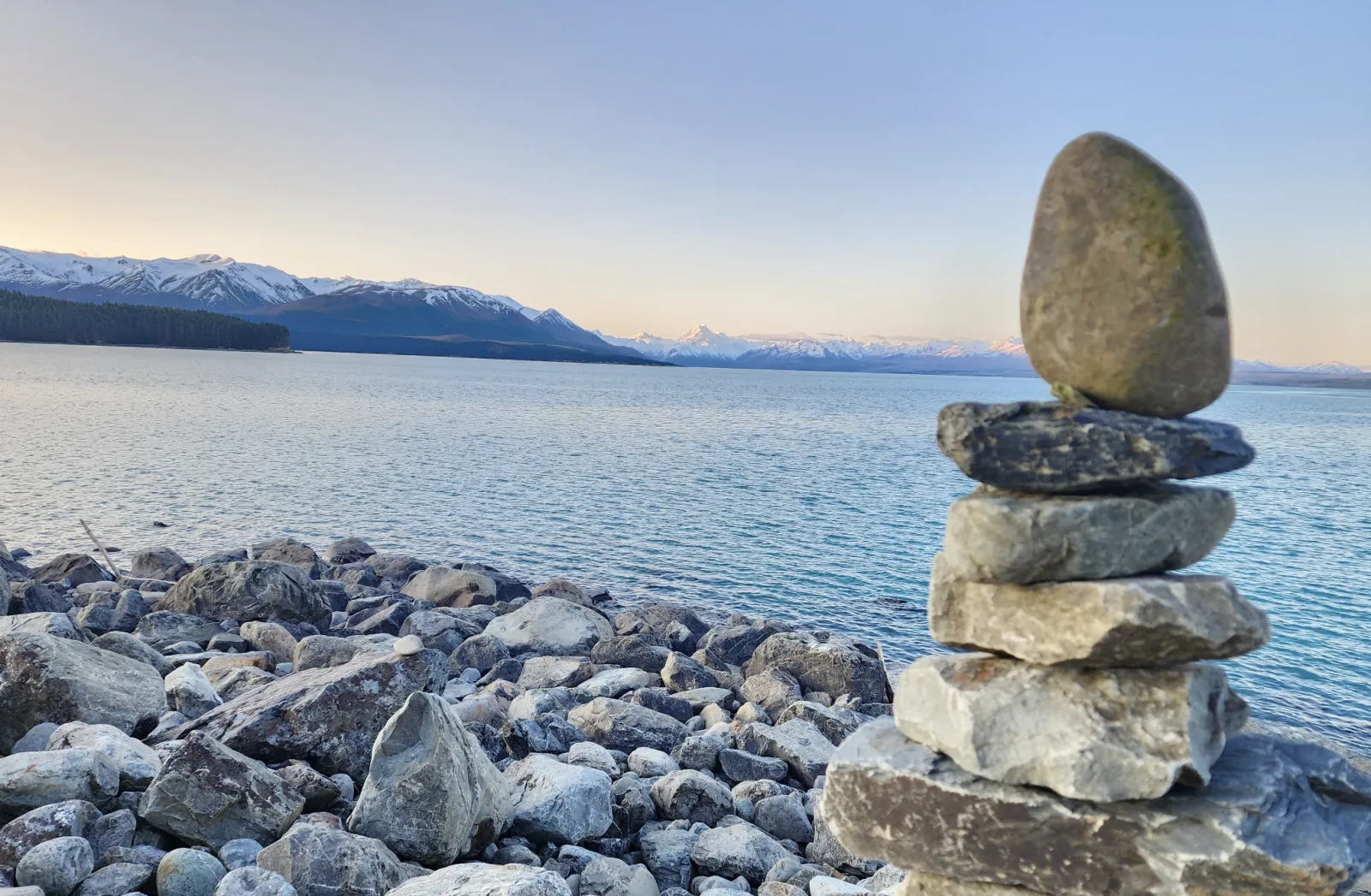 Stacked stone cairn on the Lake Pūkaki shore, with Aoraki/Mount Cook faint on the skyline at dusk