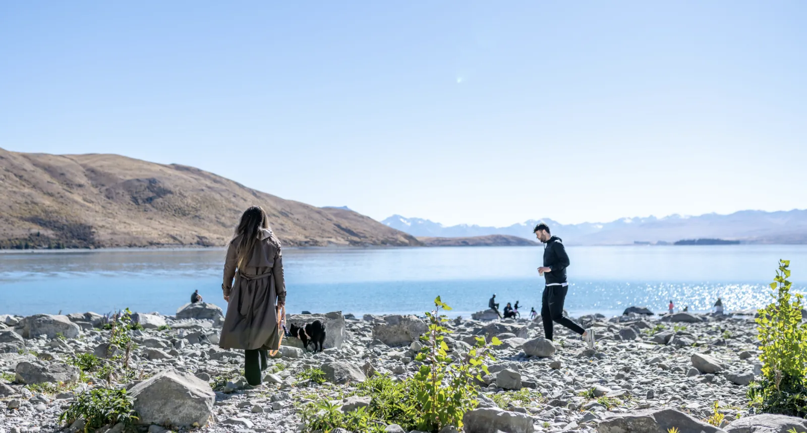 People and a dog on the stony shore of Lake Tekapo, with pale blue water and distant snowy ranges