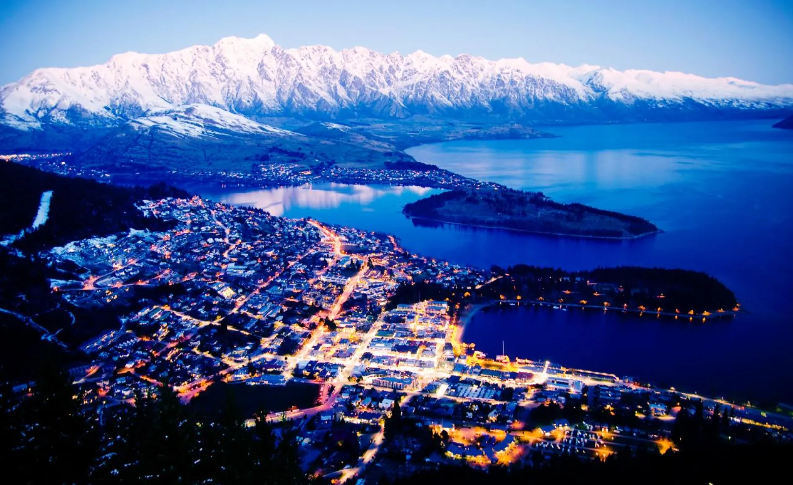 Queenstown lit up at dusk on Lake Wakatipu below The Remarkables, where the one-way tour finishes