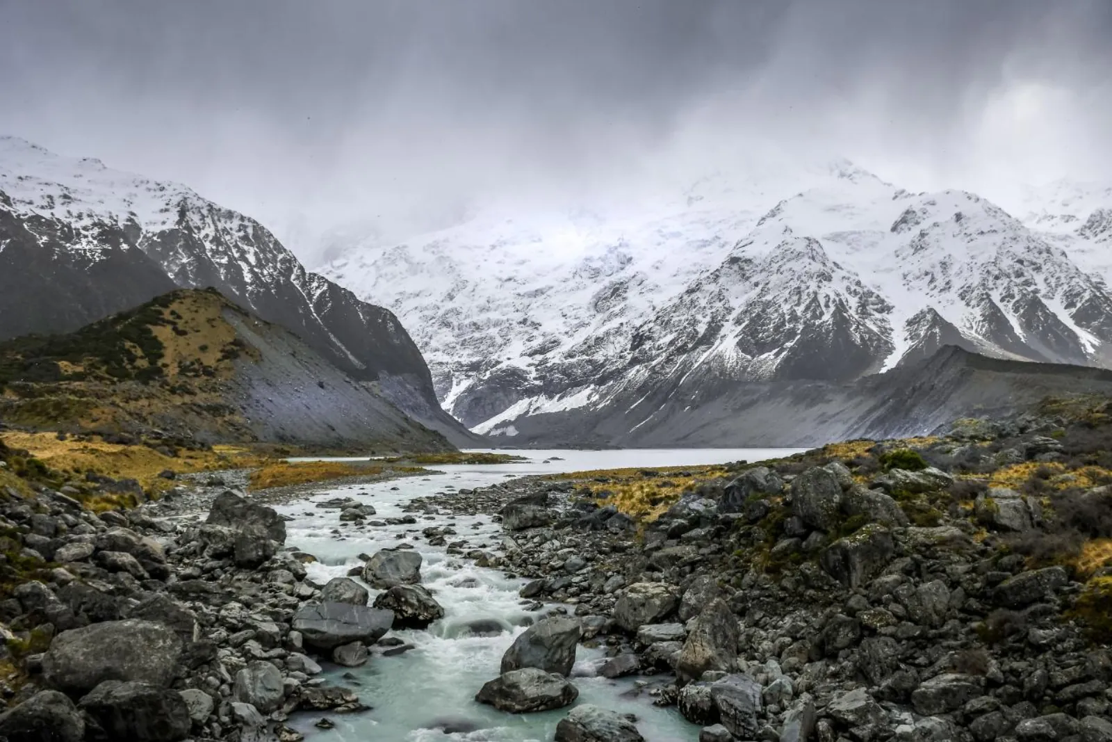 Glacial river running over boulders in the Hooker Valley toward Hooker Lake, under low snow cloud