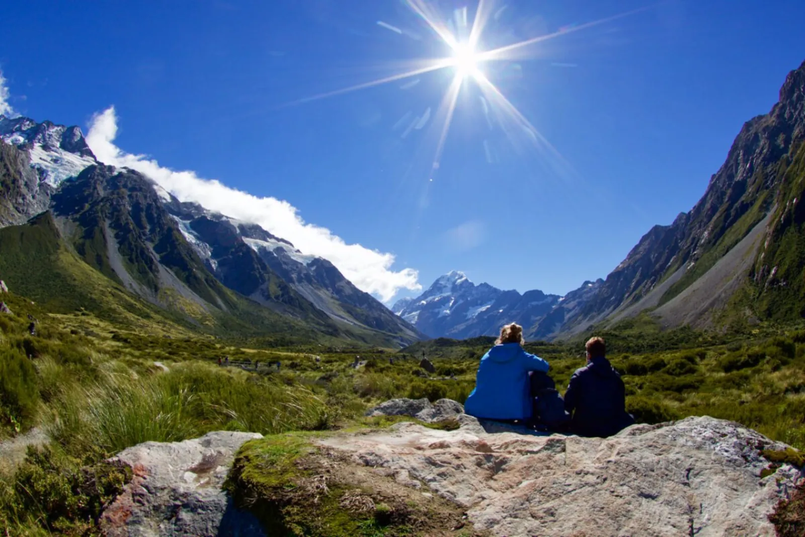 Two walkers resting on a rock in the Hooker Valley, looking toward Aoraki/Mount Cook under a bright sun