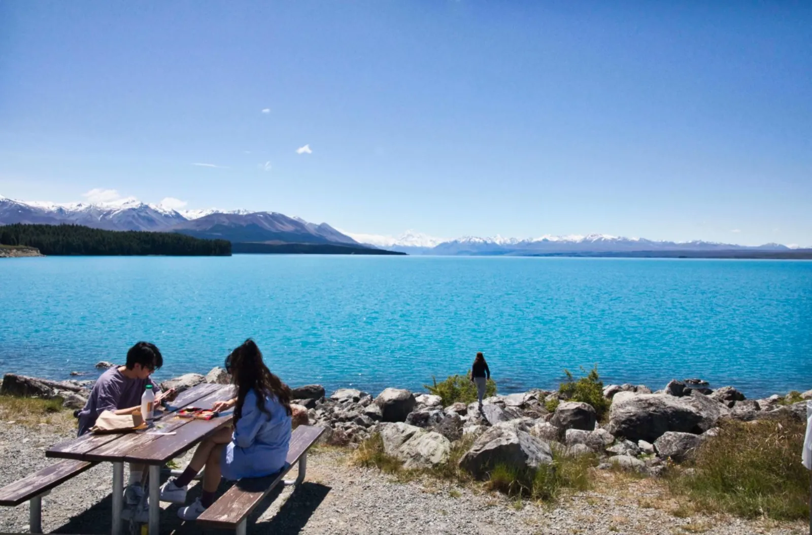 Travelers at a picnic table on the rocky shore of turquoise Lake Pūkaki, with the Southern Alps on the skyline