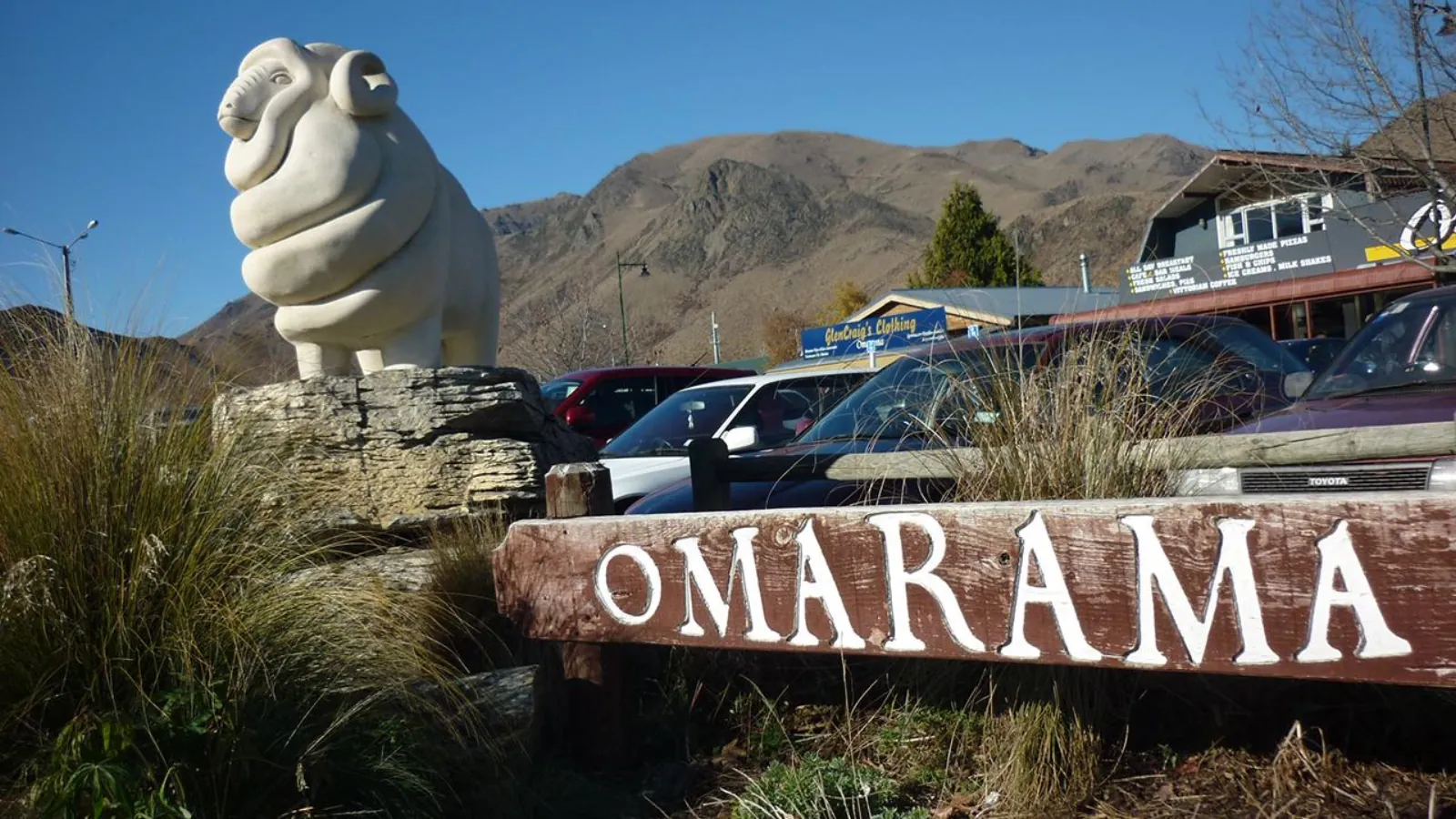 Carved stone merino ram and the wooden Omarama town sign, with cafés and brown hills behind