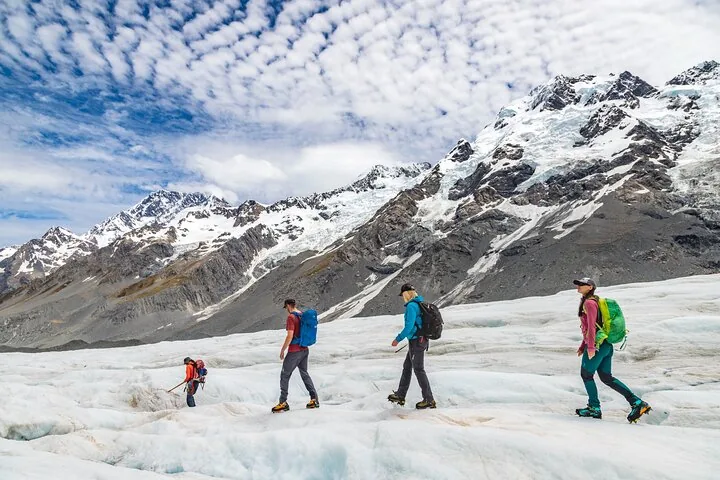 Four hikers in crampons walking across the Tasman Glacier below snowy ridges