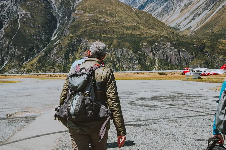 Passenger with a backpack walking across the airstrip toward a red and white plane below steep mountains