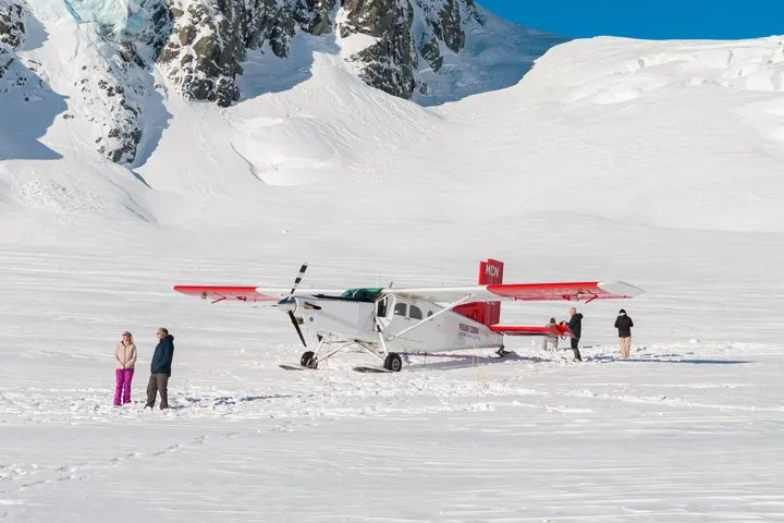 Ski plane parked on a flat glacier snowfield, passengers standing around it in the sun