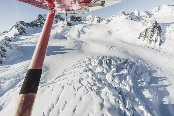 View past the ski plane's red wing strut down onto crevassed glacier ice and snowy peaks