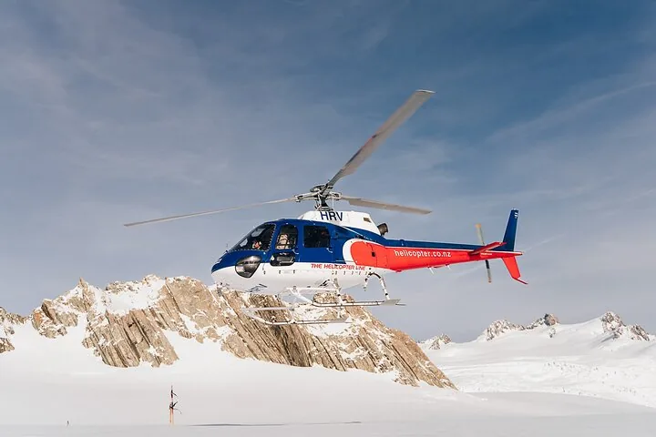 Red, white and blue helicopter hovering over a snowfield beside a rocky outcrop