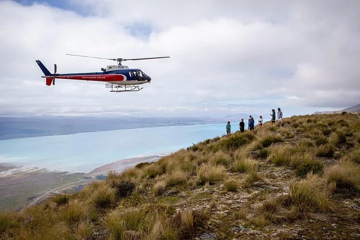Helicopter flying past hikers on a tussock ridge above the turquoise water of Lake Pūkaki