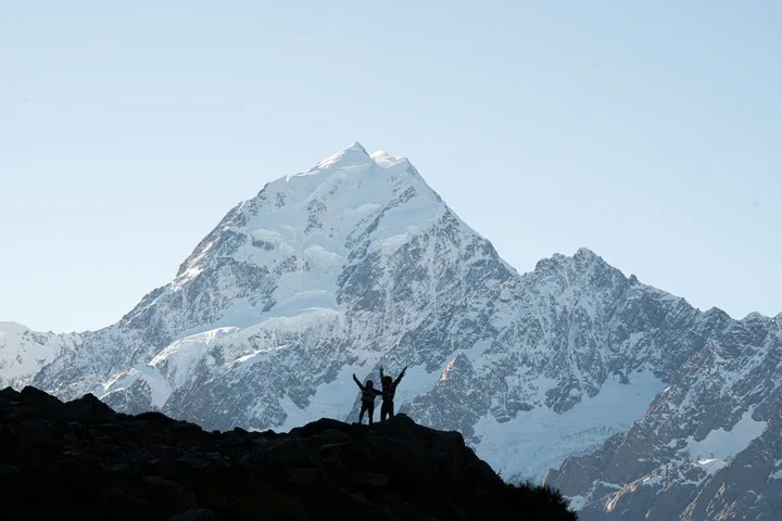 Two people silhouetted with raised arms on a ridge below the snowy summit of Aoraki/Mount Cook